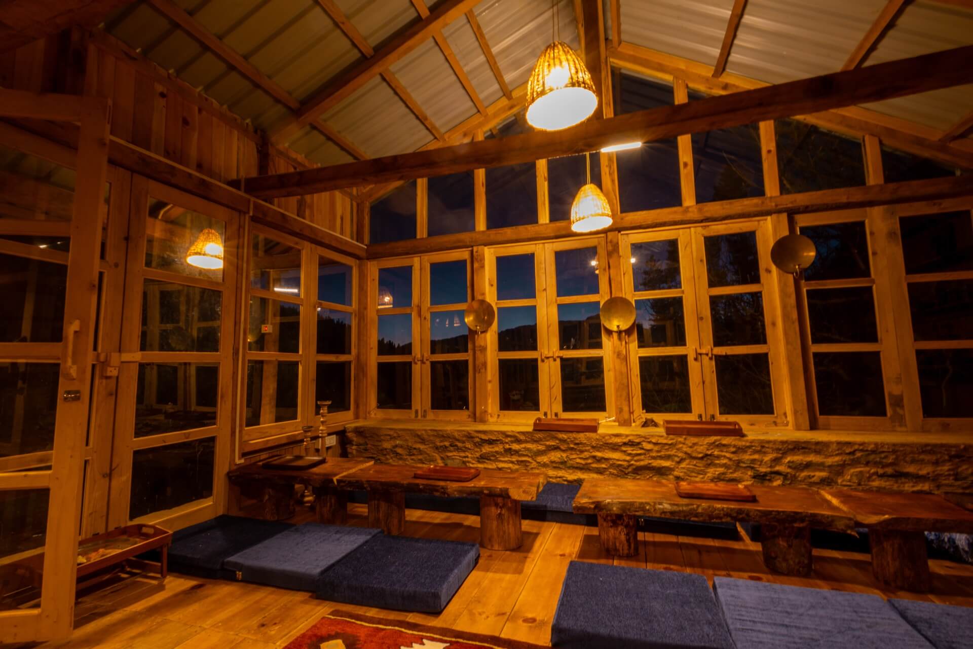 Glass-walled common room at Wild lit warm at dusk, low wooden tables and floor cushions